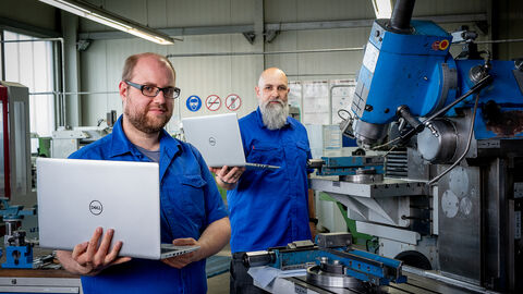 Zwei Angestellte stehen schräg hintereinander mit Laptops in der Hand an Maschinen.