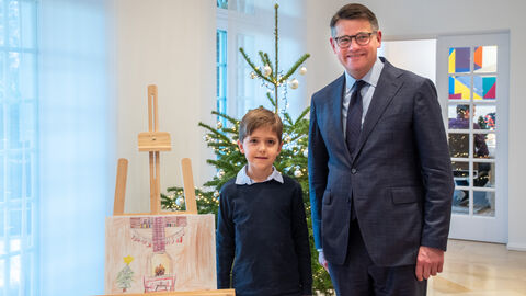 Der achtjährige Jakob steht mit Boris Rhein neben seinem gemalten Weihnachtsbild. Beide gucken in die Kamera