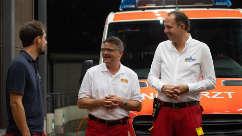 Ministerpräsident Rhein im Gespräch mit zwei Personen des ASB vor einem Einsatzfahrzeug.