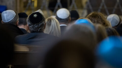Männer und Frauen sitzen auf Stühlen und sind von hinten zu sehen. Die Männer tragen eine Kippa.