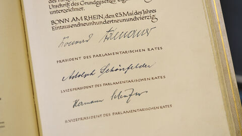 Blick auf die Urschrift des Grundgesetzes der Bundesrepublik Deutschland. Hier ist die Seite mit den Unterschriften des Präsidenten des Parlamentarischen Rates Konrad Adenauer, sowie des Ersten Vizepräsidenten des Parlamentarischen Rates Adolph Schönfelder und des Zweiten Vizepräsidenten des Parlamentarischen Rates Hermann Schäfer zu sehen.