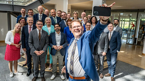 Bürgermeister Sebastian Wysocki macht ein Selfie mit dem Kabinett Über 20 Personen stehen dicht nebeneinander und posieren für ein Bild. Vorne steht ein Mann, der ein Handy in der Hand hält und von sich und den anderen ein Selfie macht.