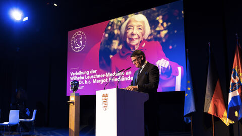 Person steht an Rednerpult. Auf dem Bildschirm im Hintergrund sieht man ein Foto einer älteren Frau.