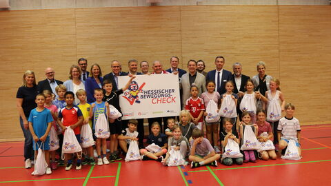 Sportminister Beuth und Kultusminister Lorz zwischen Kindern und Erwachsenen in einer Turnhalle. Sie stehen zum Gruppenfoto aufgestellt nebeneinander und halten ein Schild "Hessischer Bewegungs-Check" in den Händen.