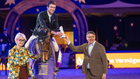 Edward Levy sitzt auf seinem Pferd und beugt sich zu Ministerpräsident Rhein herunter, der ihm eine Trohäe überreicht. Daneben steht die Veranstalterin des Festhallen Reitturniers, Ann Kathrin Linsenhoff.