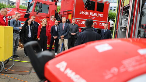 Martin Rößler und eine Gruppe von Menschen steht vor einem Feuerwehrauto. Im Vordergrund sieht man eine Person von hinten, die der Gruppe etwas erzählt.